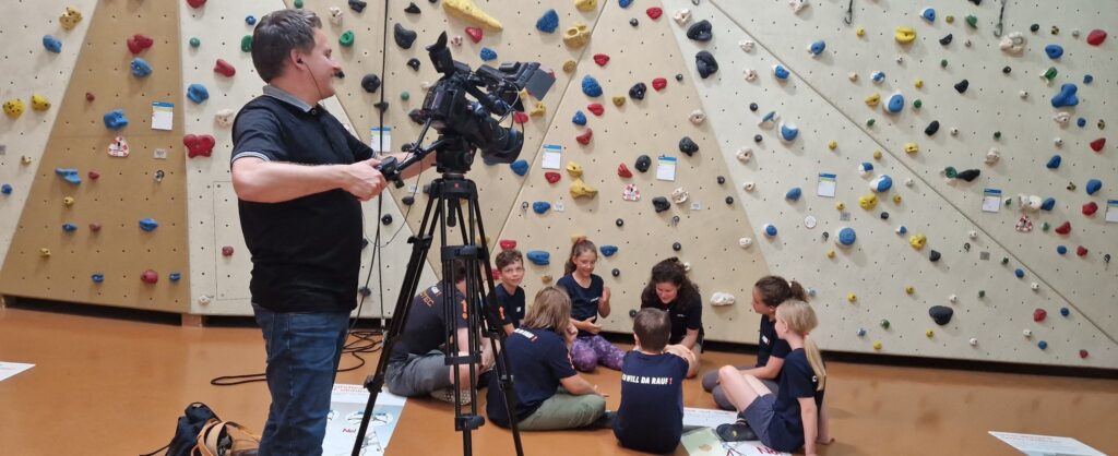 Kameramann hält Fernsehkamera auf Kinder, die im Kreis auf dem Boden sitzen. Im Hintergrund sieht man die Klettergriffa an der Wand in der Kletterhalle.