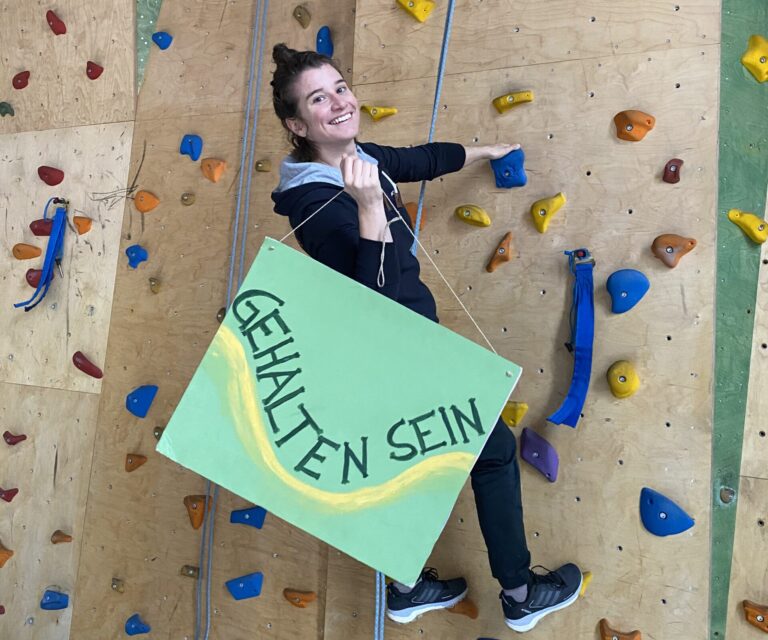 Eine Frau an der Kletterwand hält ein großes grünes Schild in einer Hand mit der Aufschrift " Gehalten sein"