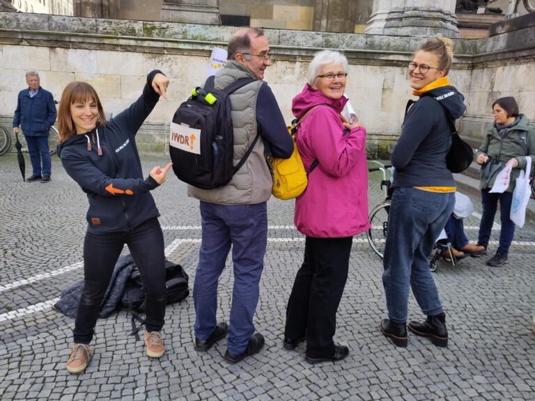 Vier Personen stehen nebeneinander. Die Frau links zeigt mit ihren Händen auf dieden Rucksack des Mannes neben rechts neben ihr. Auf dem Rucksack ist "IWDR" zu lesen. Neben dem Mann stehen noch zwei Frauen udn schauen in die Kamera