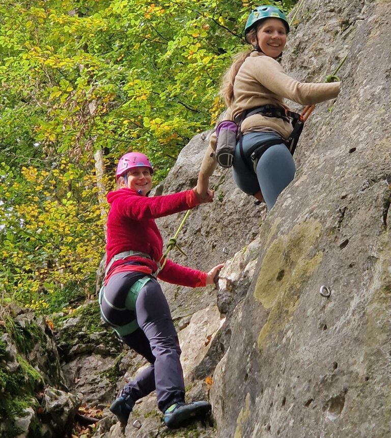 Zwei Frauen mit Schutzhelmen und Klettergurten und mit Seilen gesichert geben sich an einem Fels die Hand und lächeln in die Kamera. Von unten fotografiert