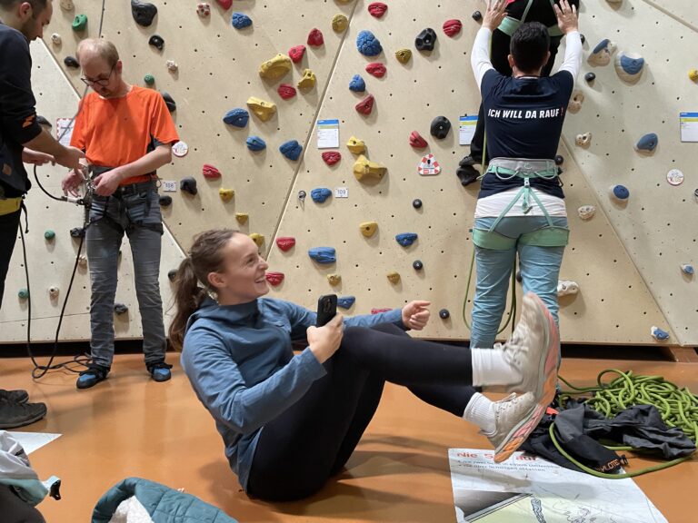 Eine Frau sitzt auf dem Boden vor einer Kletterwand in der Halle und lacht. Im Hintergrund stehen Kletternde an der Wand