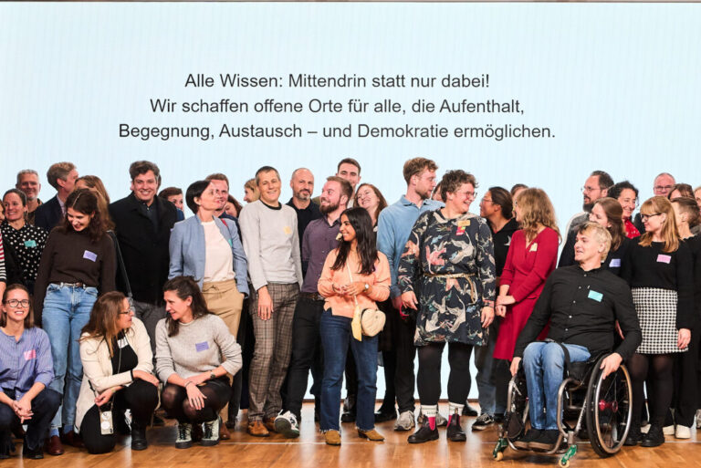 Eine Gruppe von ca 60 Personen vor einer großen Wand mit der Aufschrift "Alle wissen: Mittendrin statt nur dabei! Wir schaffen offene Orte für alle, die Aufenthalt, Begegnung, Austausch - und Demokratie ermöglichen."