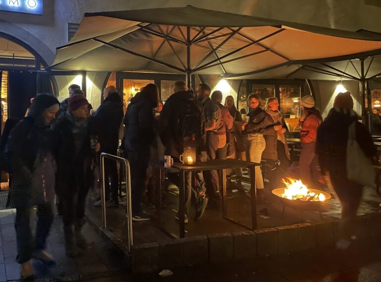 Es ist dunkel, draußen. Unter einem großen Sonnenschirm stehen eta 20 Menschen in der Dunkelheit nicht gut erkennbar. Es brennt ein Feuer in einer Schale am Boden. Im Hintergrund steht mit Leuchtschrift am Haus: "Mozzamo"