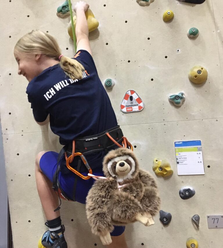 Ein Mädchen klettert an der Kletterwand und ist in Rckenansicht zu sehen. Es trägt ein T-Shirt mit der Aufschrift "Ich will da rauf!" Auf ihrem Rücken ist mit einem dünnen Gurt ein Plüsch-Faultier festgeschnallt.